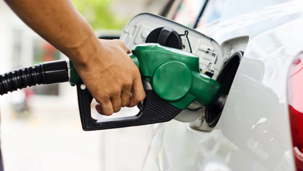 Bomba de combustível abastecendo veículo em posto de gasolina com painel de preços ao fundo, representando a alta da gasolina e do diesel no Brasil.