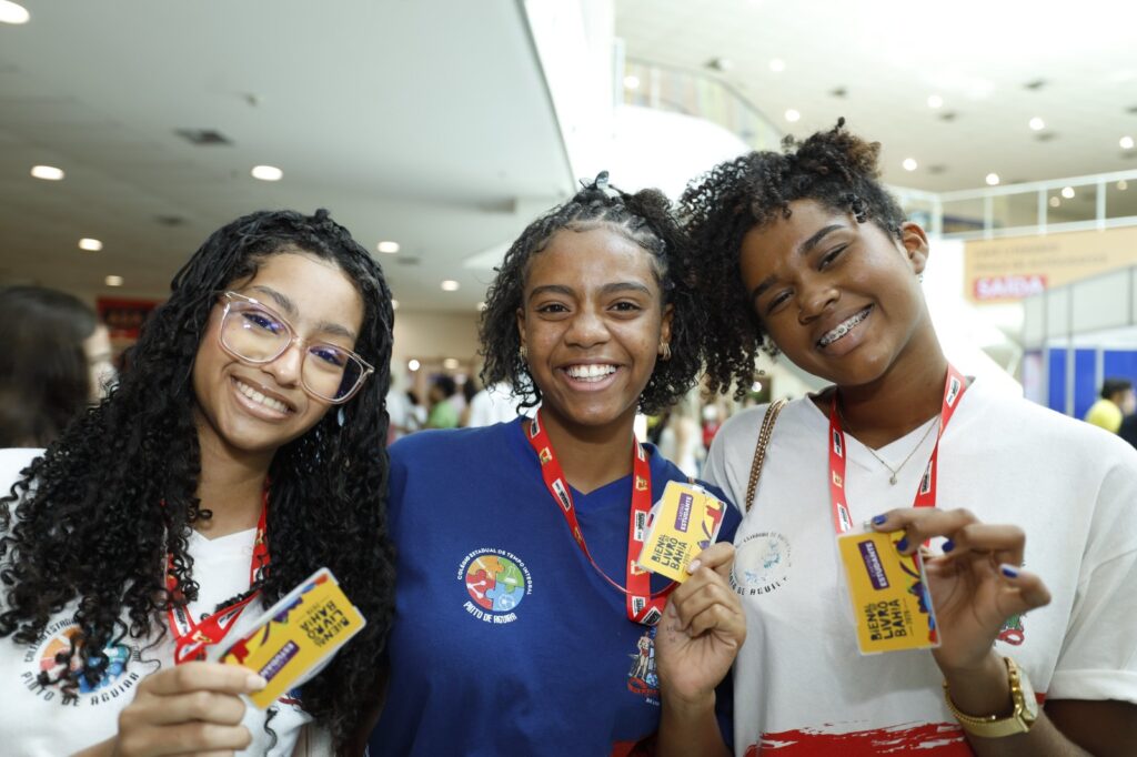 Três estudantes baianas sorrindo em ambiente escolar, representando os investimentos do Governo da Bahia em educação, leitura e cultura na campanha Do Lado da Cultura 2026.